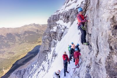 Personen mit Helmen und Kletterausrüstung an einer schneebedeckten Felswand in den Bergen.