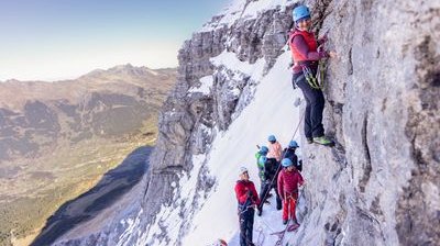 Personen mit Helmen und Kletterausrüstung an einer schneebedeckten Felswand in den Bergen.