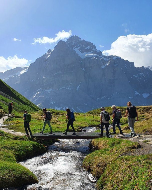 Hikers crossing a stream with a bridge with the Wetterhorn in the background.