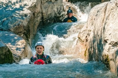 Zwei Personen beim Canyoning in einem felsigen Bach, die Helme und Neoprenanzüge tragen, umgeben von fließendem Wasser.