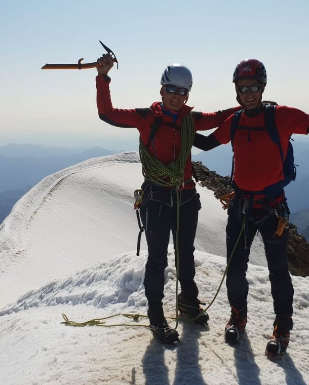 Climbers in helmets and harnesses stand on a snowy mountain ridge, holding ice axes, with others behind.