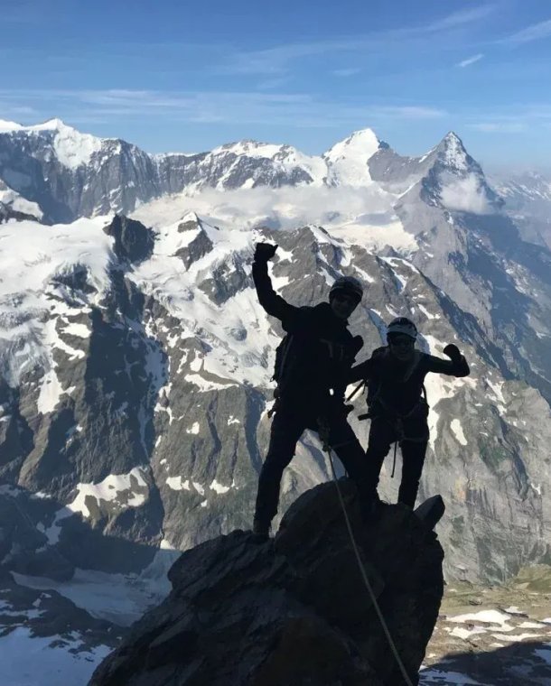 Two climbers with helmets and harnesses on a snowy mountain peak, with a vast alpine backdrop.