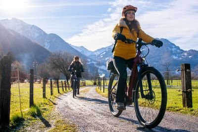Two cyclists wearing helmets ride on a gravel path with mountains in the background.
