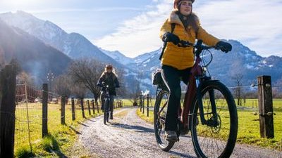 Two cyclists wearing helmets ride on a gravel path with mountains in the background.