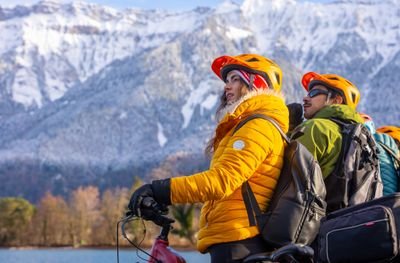 People wearing helmets and jackets on bicycles by a lake with snow-capped mountains in the background.