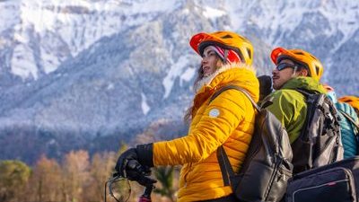 People wearing helmets and jackets on bicycles by a lake with snow-capped mountains in the background.
