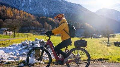 Person cycling on a paved path, wearing a helmet and backpack, with snowy mountains in the background.
