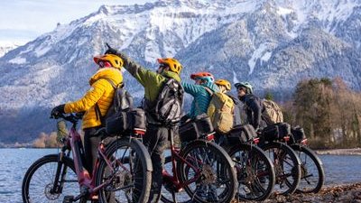 Five people on bikes with helmets and backpacks by a lake, snowy mountains in the background.