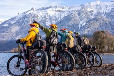 Fünf Personen auf Fahrrädern mit Helmen und Rucksäcken am See, verschneite Berge im Hintergrund.