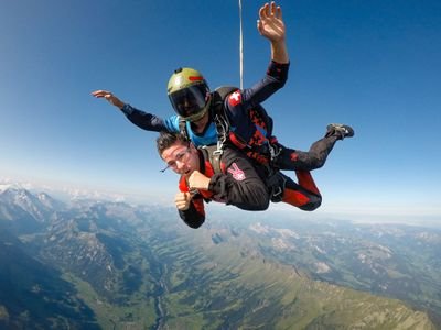 Two people tandem skydiving over mountains, wearing helmets, goggles, and harnesses.