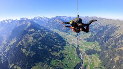 Two people tandem skydiving over a mountainous landscape, wearing helmets and harnesses.