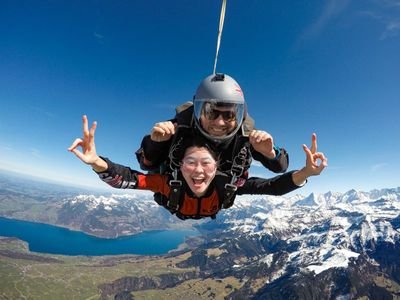 Two people tandem skydiving over snow-capped mountains and a lake, wearing helmets and harnesses.