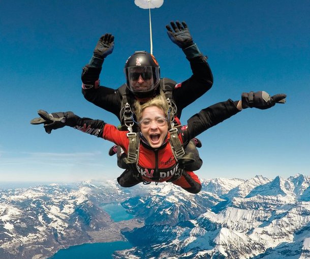 Two people tandem skydiving over snowy mountains, wearing helmets and harnesses.