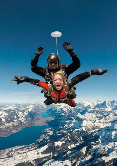 Two people tandem skydiving over snowy mountains, wearing helmets and harnesses.