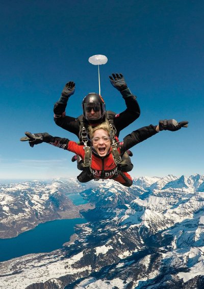 Two people tandem skydiving over snowy mountains, wearing helmets and harnesses.
