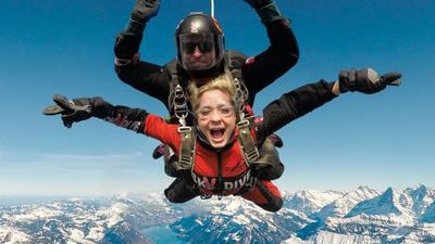 Two people tandem skydiving over snowy mountains, wearing helmets and harnesses.