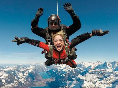 Two people tandem skydiving over snowy mountains, wearing helmets and harnesses.