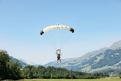 Two people paragliding with a parachute over a grassy field, mountains in the background.