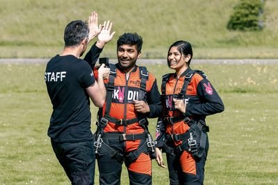 Two people in skydiving suits and harnesses stand on grass, interacting with a staff member.