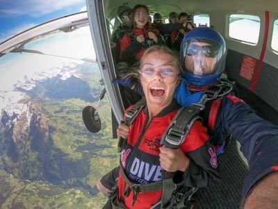 Zwei Personen in Fallschirmausrüstung, einschließlich Helmen und Gurten, bereiten sich darauf vor, aus einem Flugzeug über den Bergen zu springen.