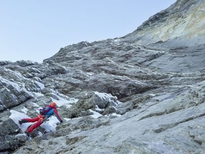 Kletterer mit Helm und Klettergurt auf felsigem Berggelände mit Schnee.