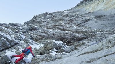 Kletterer mit Helm und Klettergurt auf felsigem Berggelände mit Schnee.
