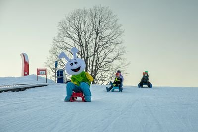 Drei Personen rodeln im Schnee, eine Person im Hasenkostüm, die anderen in Helmen und Winterkleidung.