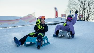 Zwei Personen rodeln im Schnee, tragen Helme, Schutzbrillen und Winterkleidung, mit Bäumen im Hintergrund.