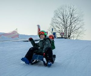 Two people sledding on snow, wearing helmets and winter gear, with a snowy hill and bare tree.