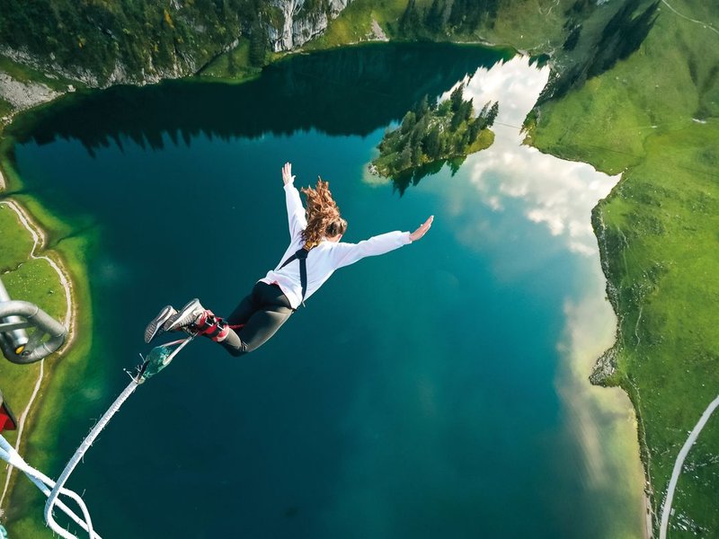 Person bungee jumping over a alpine lake, wearing a harness and ankle straps, surrounded by mountains.