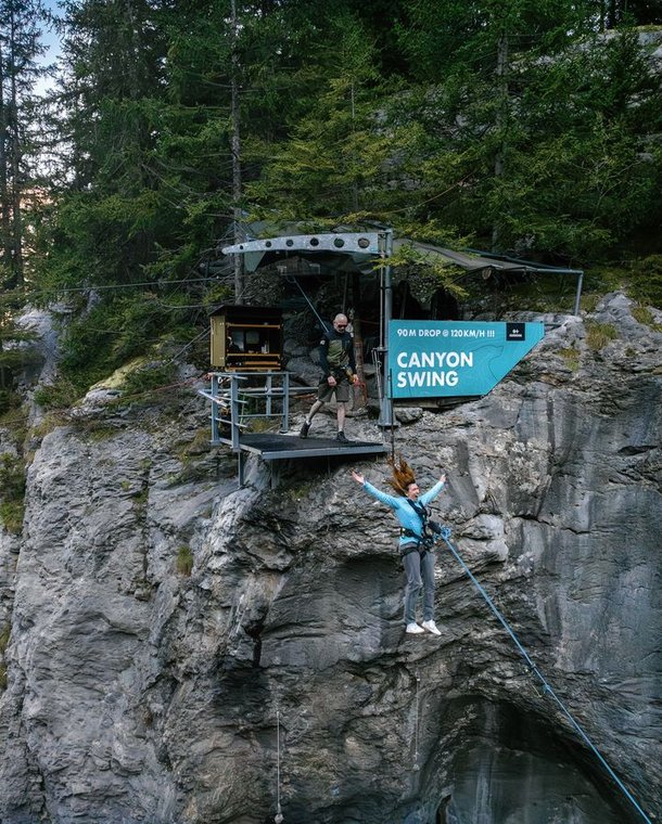 Person in harness jumping from platform for canyon swing, with guide nearby, set in rocky mountain area.