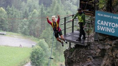 Person in harness jumps from platform for canyon swing; guide stands nearby; forested backdrop.
