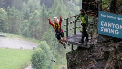 Person in harness jumps from platform for canyon swing; guide stands nearby; forested backdrop.