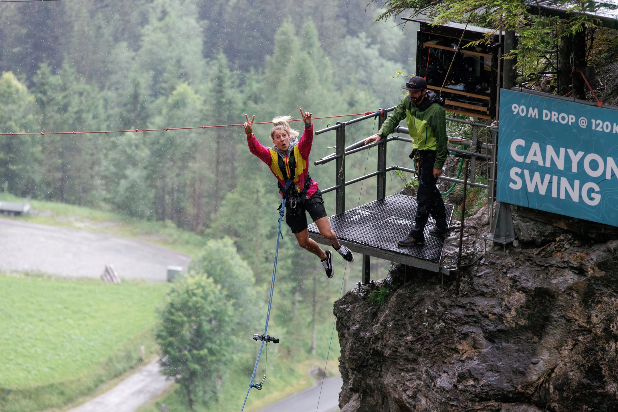Person in harness jumps from platform for canyon swing; guide stands nearby; forested backdrop.