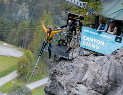 Person jumps from platform during Canyon Swing, 2 spectators, wooded canyon surroundings.