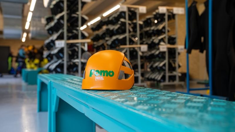 Orange climbing helmet with the inscription "Fomo" on a blue metal bench in an equipment room with shelves full of shoes.