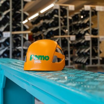 Orange climbing helmet with the inscription "Fomo" on a blue metal bench in an equipment room with shelves full of shoes.