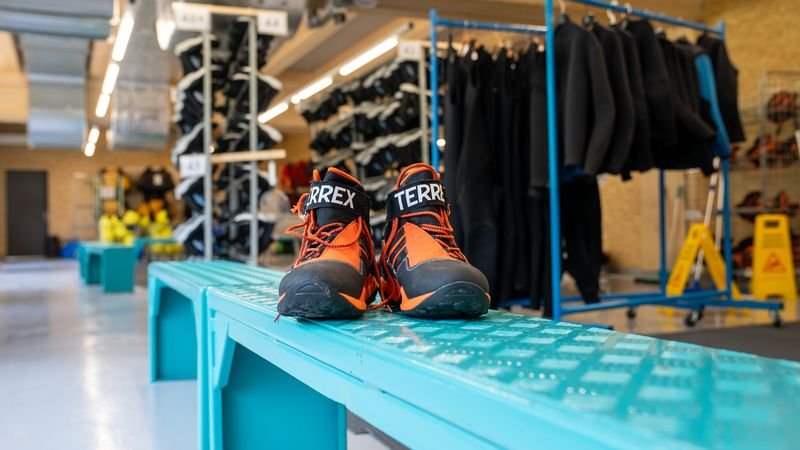 A pair of orange and black hiking shoes is standing on a blue bench in a rental room with wetsuits and equipment in the background.