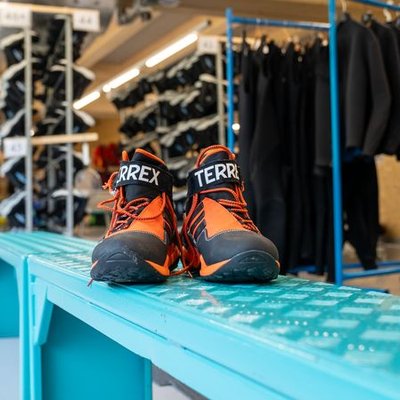 A pair of orange and black hiking shoes is standing on a blue bench in a rental room with wetsuits and equipment in the background.