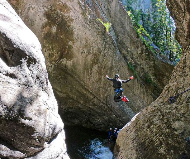 Person in wetsuit and helmet jumps into canyon pool, others watch below, surrounded by rocky cliffs.