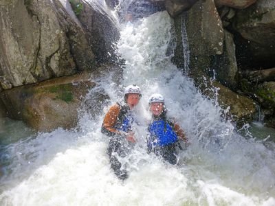 Zwei Personen in Helmen und Neoprenanzügen stehen unter einem Wasserfall in einer felsigen Canyon-Umgebung.