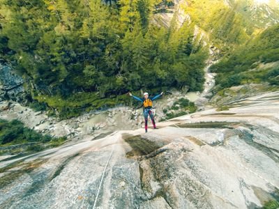 Person, der an einer steilen Felswand in einem bewaldeten Canyon abseilt, mit Helm und Klettergurt.