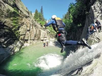 Person im Neoprenanzug und Helm springt in einen Canyon-Pool, andere warten auf den Felsen; üppige, felsige Umgebung.