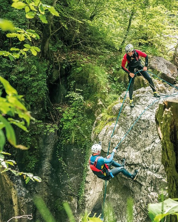 Two people canyoning with helmets and harnesses on a rocky cliff in a lush forest setting.
