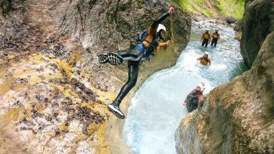 Person in wetsuit and helmet jumps into canyon pool; others in helmets and gear watch from water.