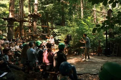 Personen mit Helmen und Kletterausrüstung hören einer Person im Wald vor Hochseilgarten zu.