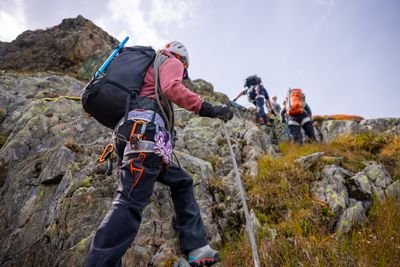 Mehrere Personen mit Helmen, Klettergurten und Rucksäcken steigen an einem Fels mit Seilsicherung in den Bergen auf.