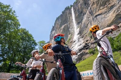 Vier Personen mit Helmen stehen mit E-Mountainbikes vor einem Wasserfall in bergiger Landschaft.