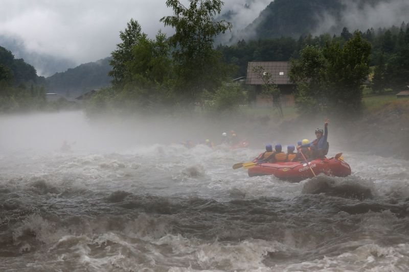 Mehrere Personen mit Helmen und Schwimmwesten beim Wildwasser-Rafting auf einem reißenden Fluss in nebliger Berglandschaft.