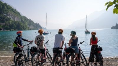 Five people with helmets stand by bikes, overlooking a lake with sailboats and mountain backdrop.